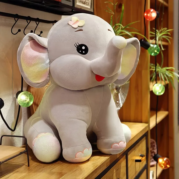 Um grande elefante cinzento de peluche com orelhas coloridas e uma flor na cabeça, sentado numa prateleira de madeira rodeada de luzes decorativas.