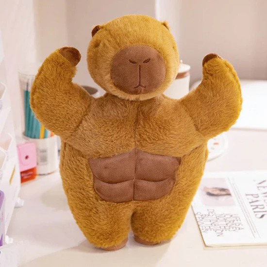 A brown plush bear toy with exaggerated muscular arms and visible abs, standing upright on a desk next to magazines and office supplies.