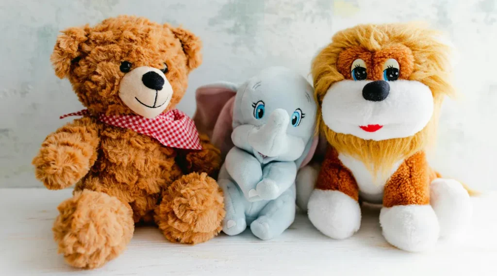 Brown bear, blue elephant, and orange lion plush toys sitting side by side on white surface.