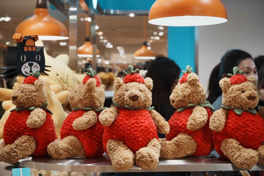 Row of cute teddy bear plush toys dressed in red strawberry outfits displayed on a shelf in a toy store
