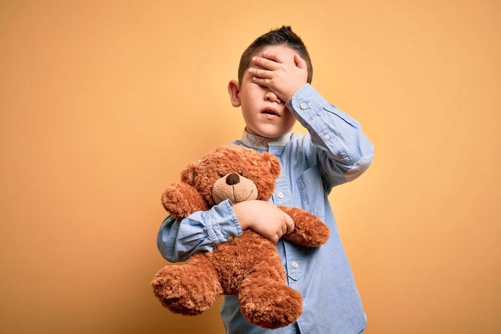 Upset boy covering face while hugging brown teddy bear against orange background