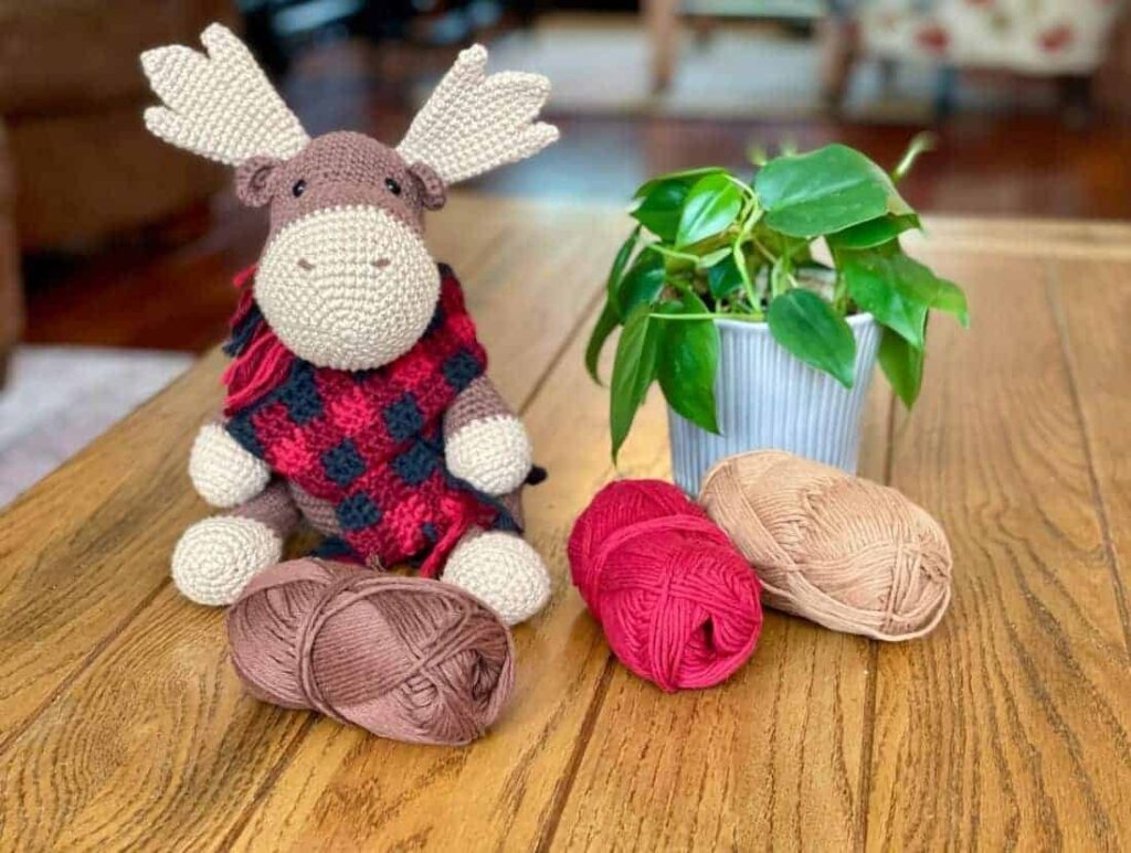 Schattige gehaakte eland knuffel met een rode en zwarte sjaal, geplaatst naast garen ballen en een groene potplant op een houten tafel