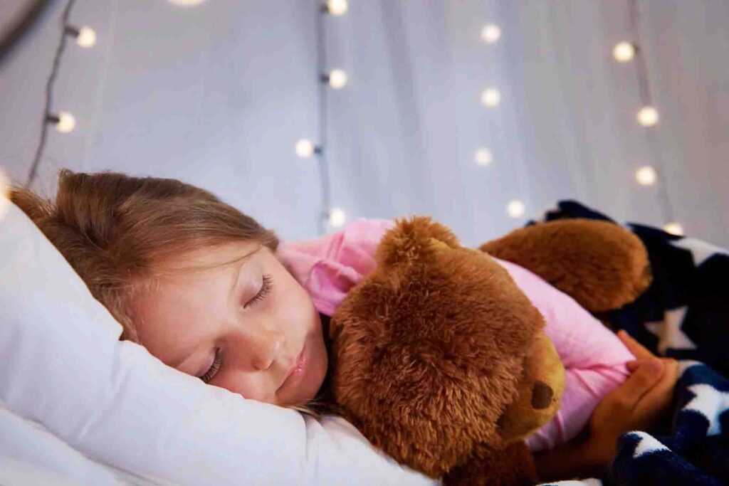Little girl peacefully sleeping while hugging a plush teddy bear under string lights in a cozy bed