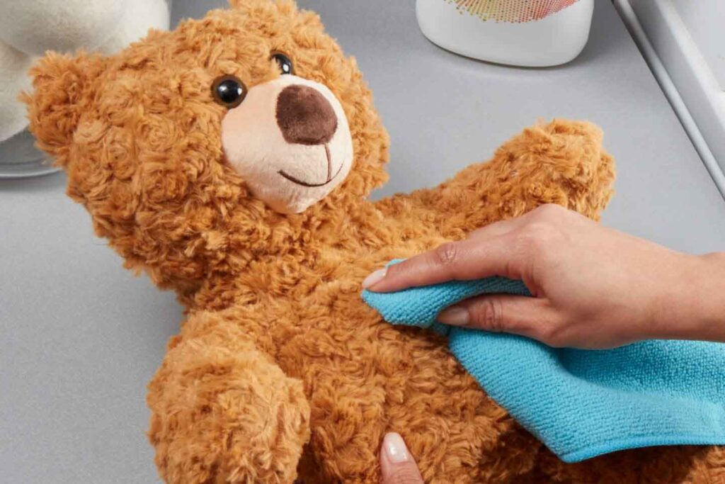 Hand using a blue microfiber cloth to clean a brown plush teddy bear on a countertop with cleaning supplies nearby