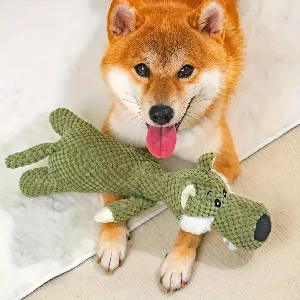 Ένας χαρούμενος σκύλος Shiba Inu ξαπλωμένος σε μια μαλακή επιφάνεια με τη γλώσσα του έξω, κρατώντας ένα πράσινο βελούδινο παιχνίδι αλιγάτορα, σχεδιασμένο για μάσημα και αγκαλιές.