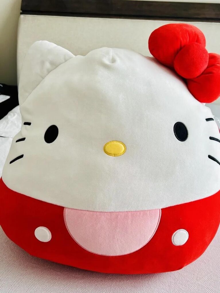 Χαριτωμένο βελούδινο μαξιλάρι Hello Kitty με κόκκινο φιόγκο σε ένα κρεβάτι.