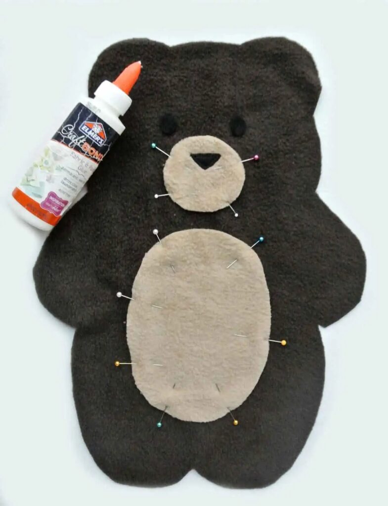 Um urso de peluche de feltro plano com alfinetes a prender a cara e a barriga bege, junto a um frasco de cola Elmer.
