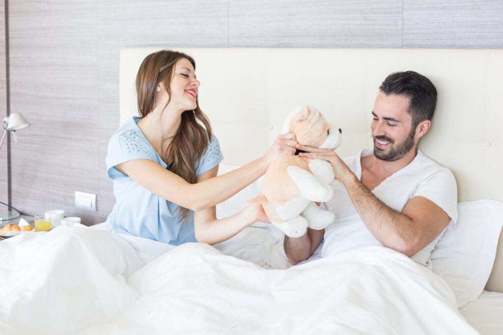 Smiling couple sitting in bed playfully holding a plush dog toy in a bright, cozy bedroom