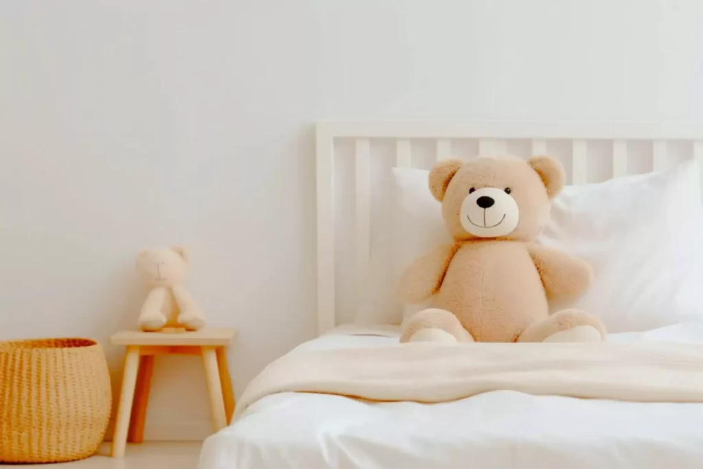 Soft beige teddy bear sitting on a white bed in a minimalistic room with another plush bear on a wooden stool
