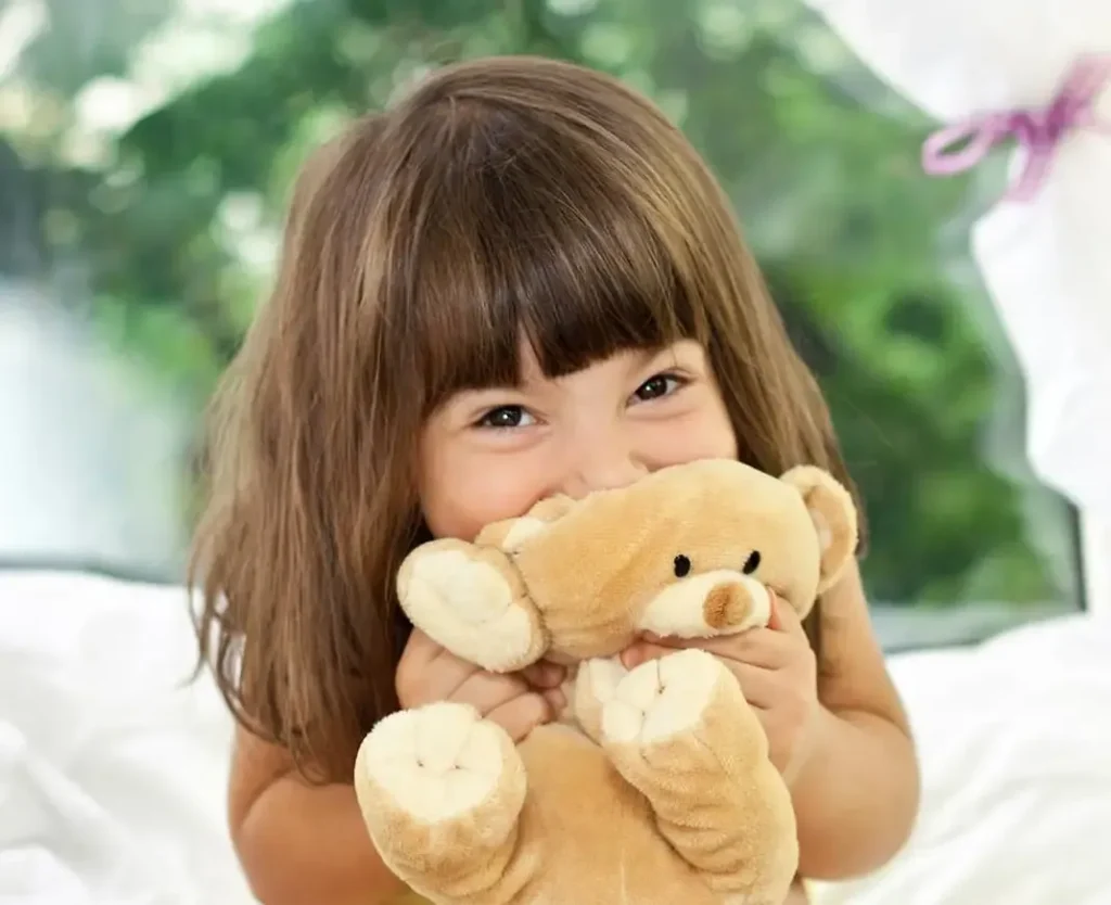 Smiling girl cuddling a soft brown teddy bear near window.