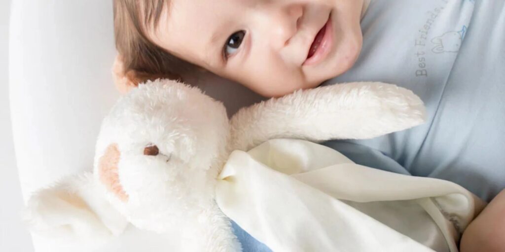 Smiling baby lying in bed cuddling a white plush bunny toy with a soft blanket