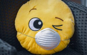 A round yellow plush pillow shaped like a winking emoji, featuring a large embroidered eye and a sewn-on white face mask, placed on a black wicker chair.