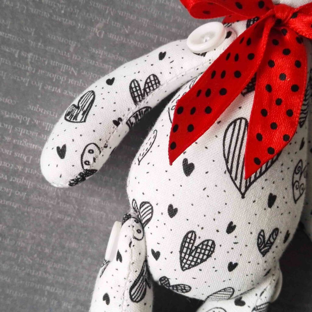 Grande plano de um peluche de tecido branco decorado com padrões de corações pretos e uma fita de bolinhas vermelhas à volta do pescoço.