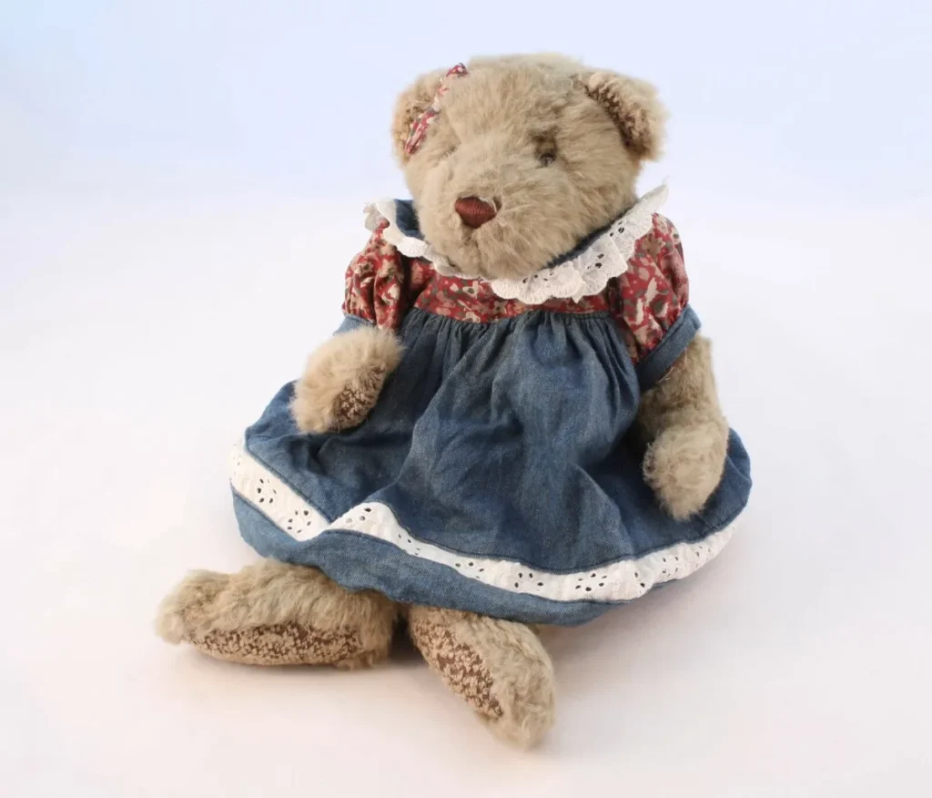 Vintage teddy bear wearing blue dress with floral pattern sitting on white background.