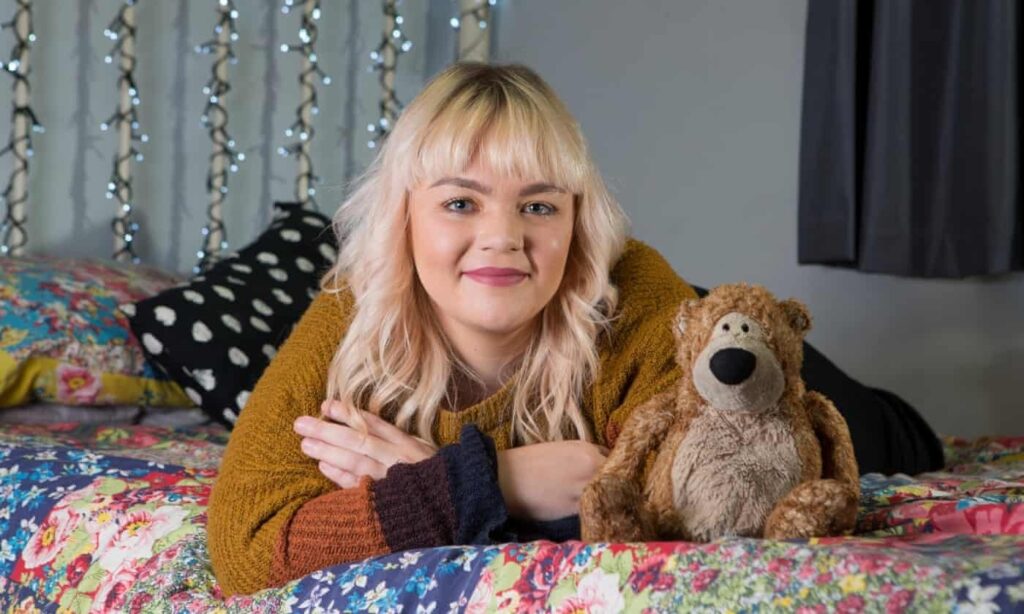 Smiling woman lying on a colorful bed next to a well-loved plush teddy bear, with fairy lights in the background