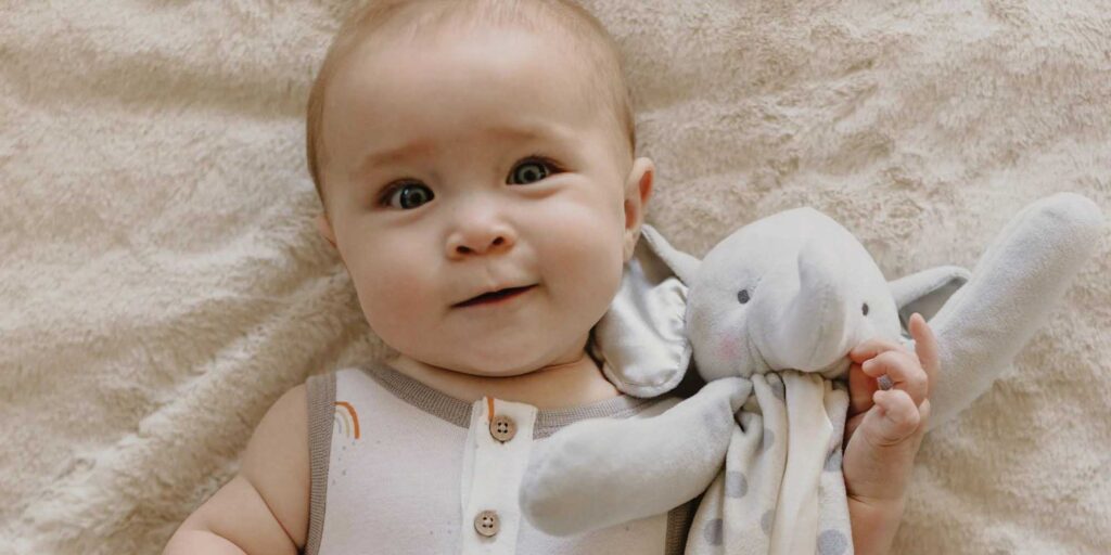 Adorable baby lying on soft beige blanket holding a grey plush elephant toy, happy infant with curious expression, cuddly stuffed animal with polka-dot fabric, cozy and comforting playtime moment, soft lighting in nursery setting.