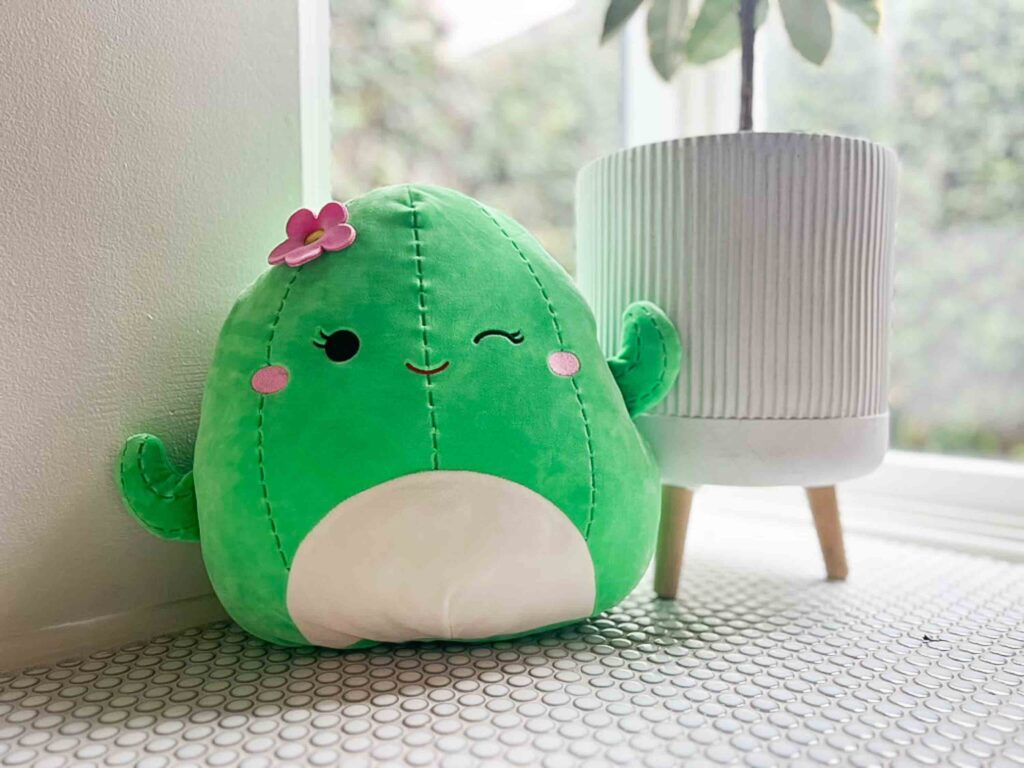 Cute green cactus plush toy with a pink flower and winking face sitting on a tiled windowsill beside a white ribbed plant pot on wooden legs, cozy and bright indoor setting with natural light.