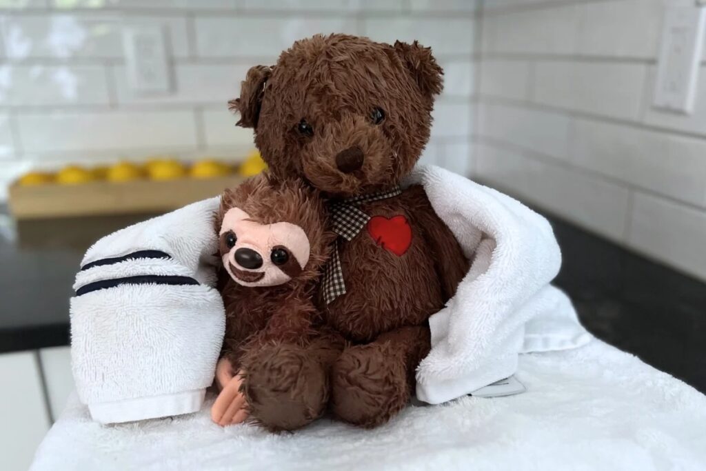 Brown teddy bear with red heart and small sloth plush wrapped in white towel on countertop.