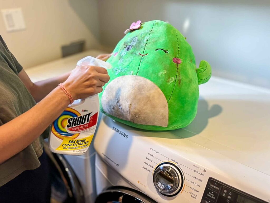 Person using Shout stain remover spray on a dirty green plush cactus toy sitting on top of a Samsung washing machine, stained stuffed animal with pink flower and smiling face, preparing for cleaning process.