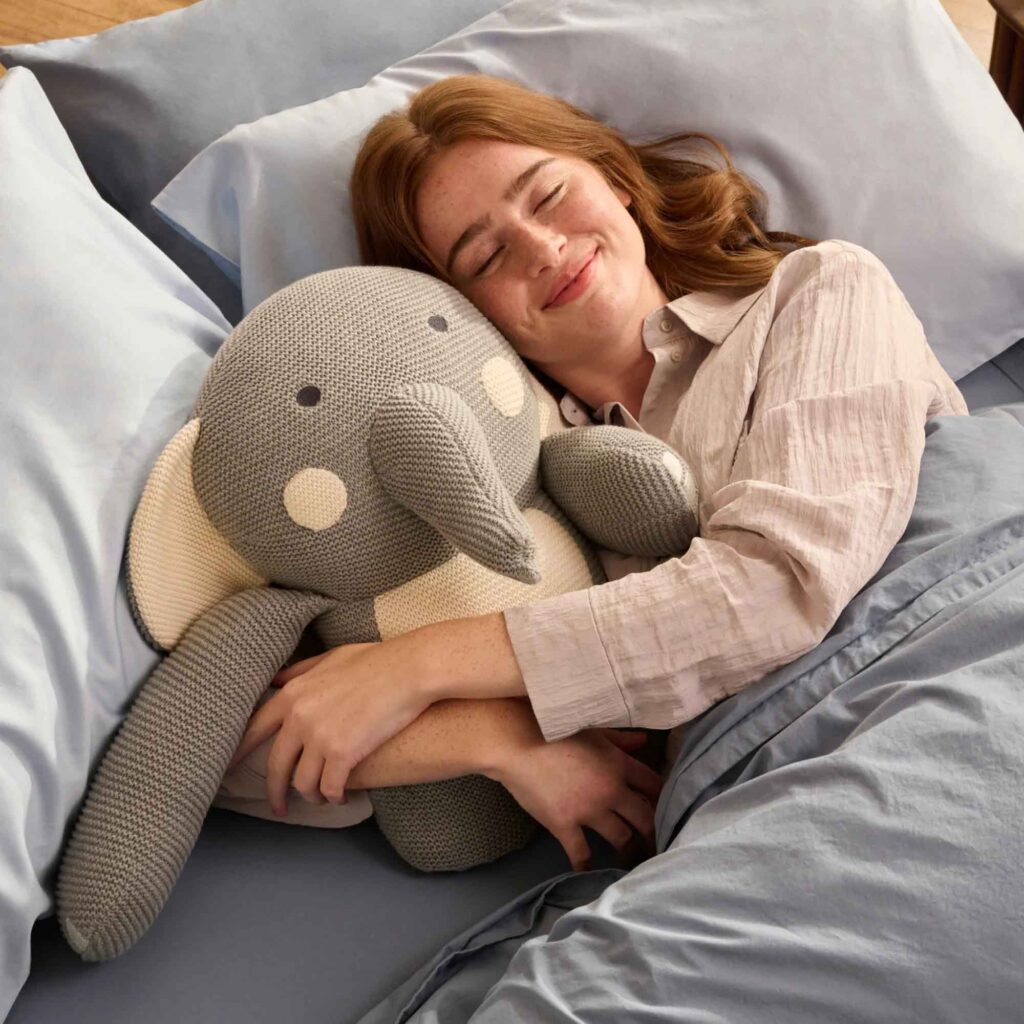 Woman happily cuddling a large knitted elephant plush toy in bed, cozy sleep environment with blue sheets and pillows, soft handmade stuffed animal, relaxing bedtime moment, comforting toy companion, peaceful expression, cute elephant design.