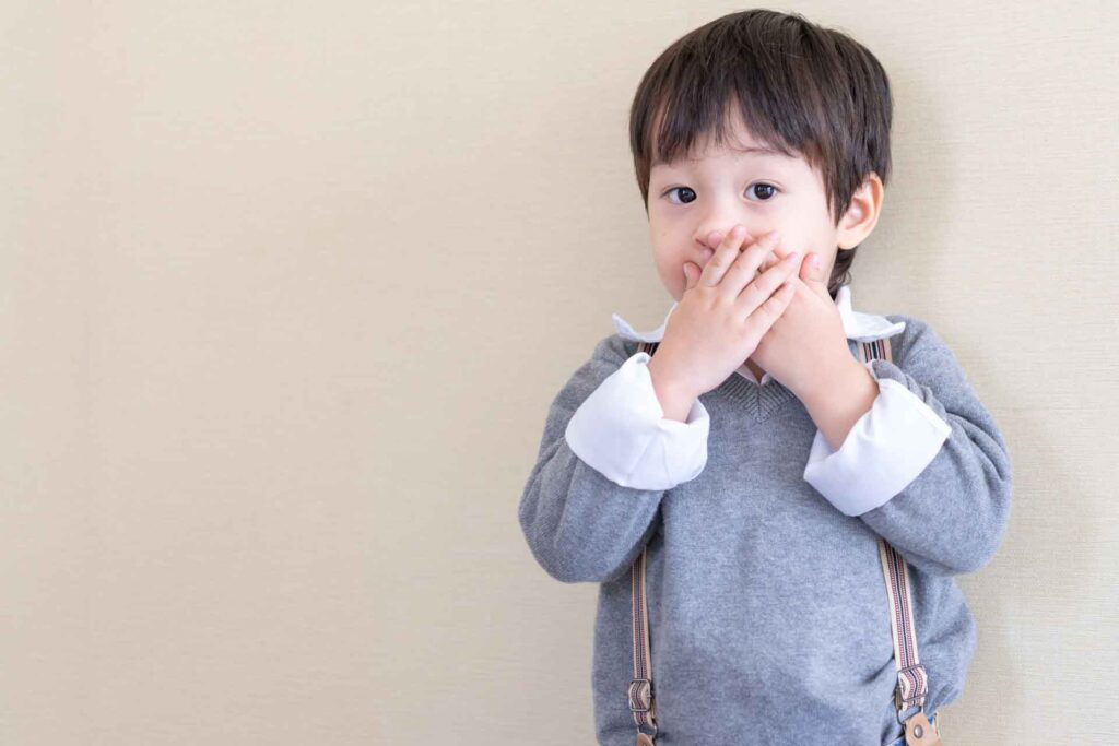 Young boy in a gray sweater and suspenders covering his mouth with both hands, surprised or playful expression, standing against a light beige wall, wearing white collared shirt underneath, indoor portrait of a child.