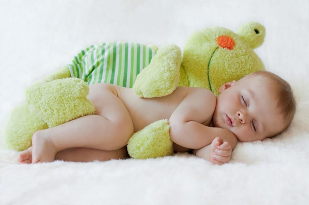 Peacefully sleeping baby snuggled up with a soft green stuffed animal that has a striped body and orange nose, lying on a fluffy white blanket.