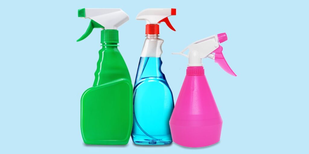 Three colorful spray bottles on a blue background, green bottle with white and green nozzle, clear bottle with blue liquid and red nozzle, pink bottle with white and pink nozzle, cleaning or disinfectant containers.