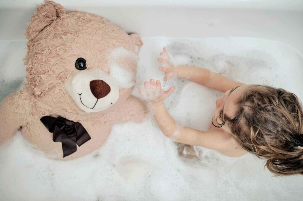 Child playing in a bubble-filled bathtub with a large plush teddy bear, both covered in soap suds, soft toy wearing a dark ribbon, fun and playful bath time moment, cozy and gentle cleaning scene.