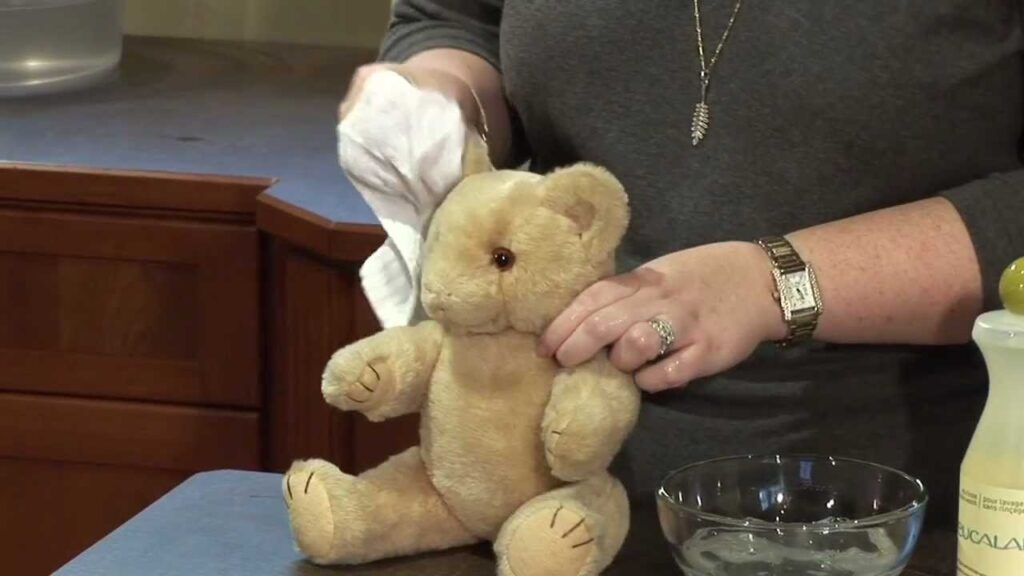 Person cleaning a beige teddy bear with a white cloth using a bowl of water and gentle detergent on a counter.