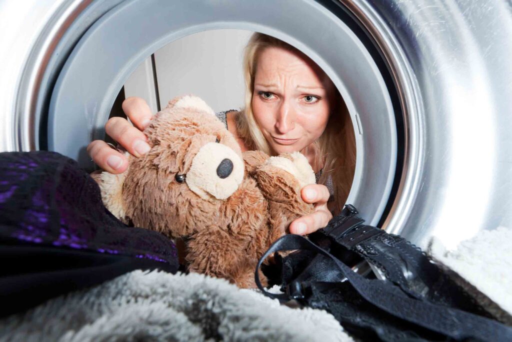 Woman with a concerned expression retrieving a worn-out teddy bear from inside a washing machine, surrounded by laundry and looking directly into the drum.