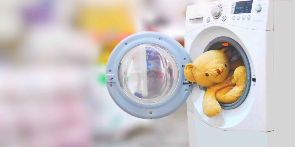 Large yellow teddy bear inside a front-loading washing machine with the door open, plush toy positioned for cleaning, home laundry appliance with control panel and dials visible, soft toy wash routine.