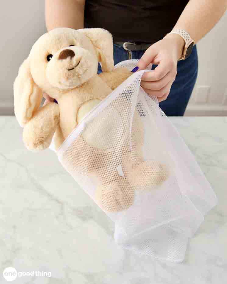 Person placing a plush dog toy into a white mesh laundry bag, preparation for washing stuffed animal safely, gentle cleaning method for soft toys, toy care routine on marble countertop, protecting plush during machine wash.