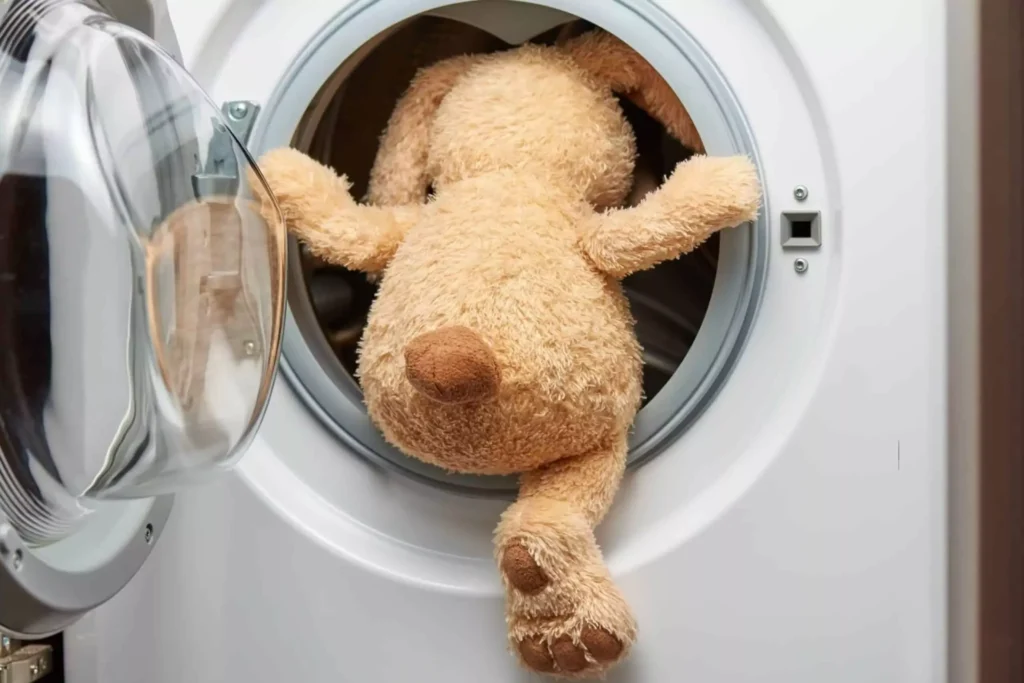 Stuffed dog toy halfway inside a front-loading washing machine with its legs hanging out and door open.