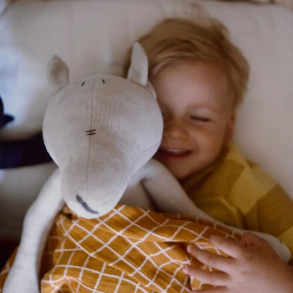 A happy child in a yellow shirt snuggles in bed with a large light gray wolf plush toy, covered by a mustard-yellow checkered blanket.