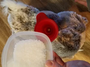 Hand holding container of white plastic stuffing beads with red funnel over plush toy opening