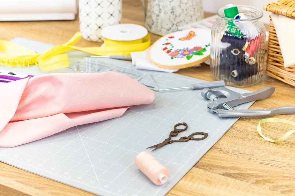 Scissors, fabric, thread, and crafting tools on cutting mat on wooden table.