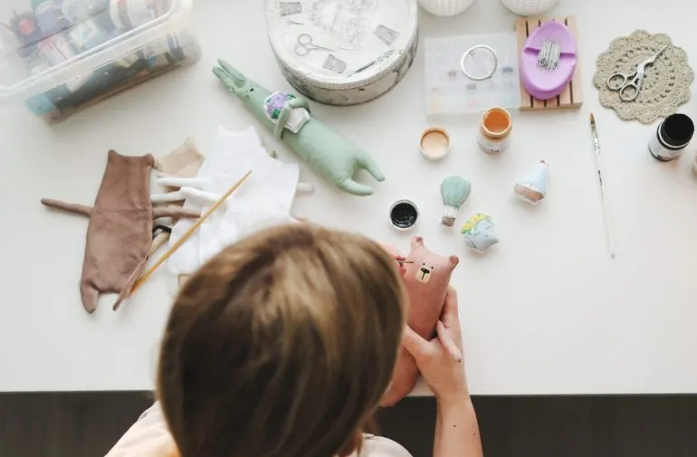 Person painting and assembling small stuffed dolls with craft supplies on table.