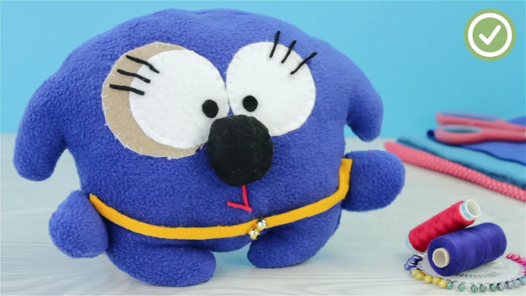 A close-up of a handmade purple stuffed animal with big eyes and a yellow belt, surrounded by crafting supplies.