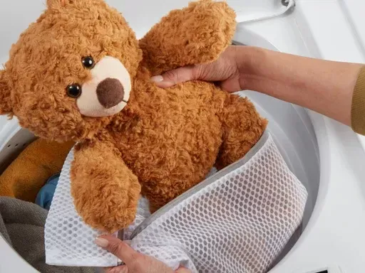 Hand placing teddy bear inside mesh laundry bag in washing machine.