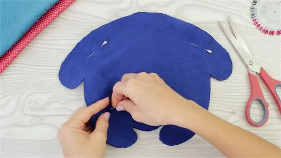A person pins fabric pieces together to form the body of a purple stuffed animal, with crafting supplies nearby.