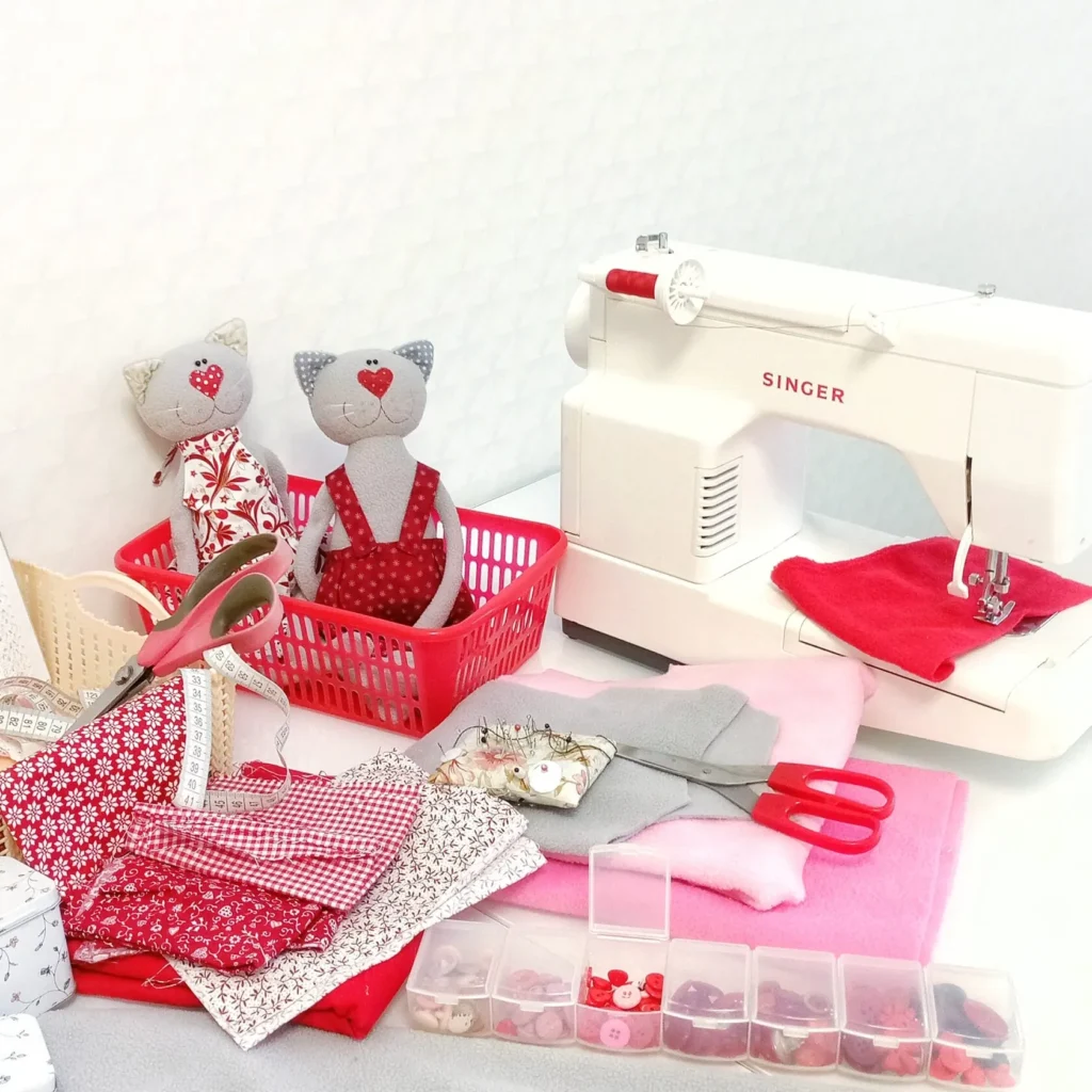 Sewing machine, scissors, measuring tape, red fabrics, buttons, pins, and two handmade plush cats in a basket on a table