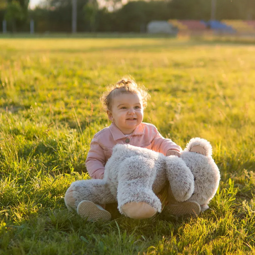 Smiling baby sitting on grass holding a large gray teddy bear outdoors.
