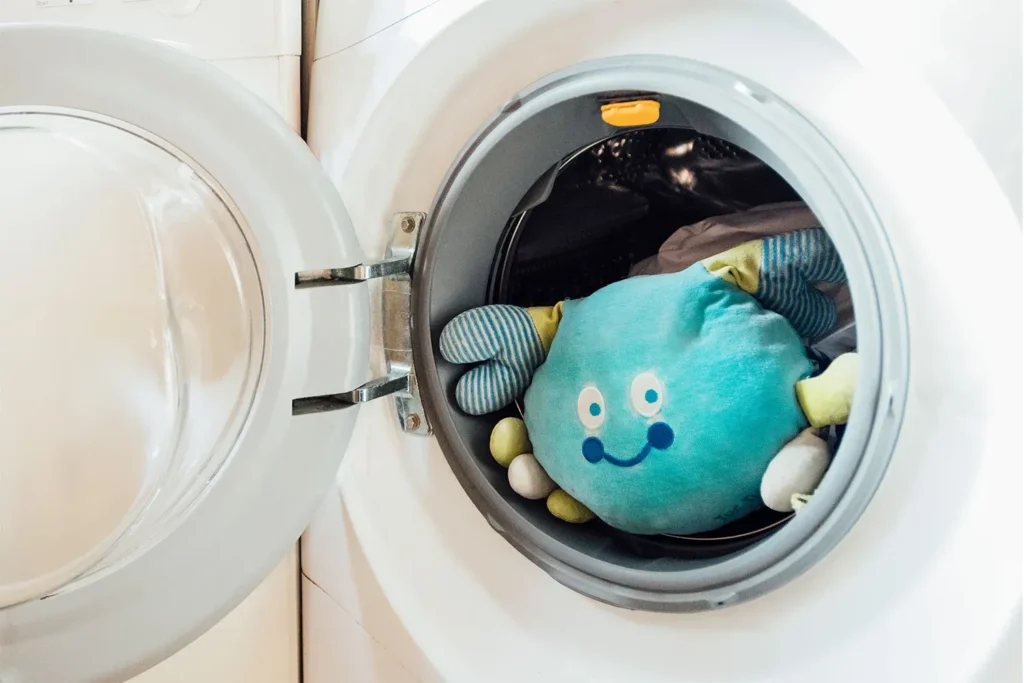 A blue smiling plush toy inside the drum of a front-loading washing machine, ready for cleaning.