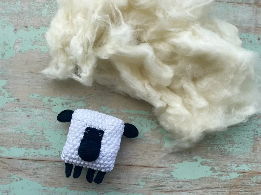 White natural wool fibers next to small crocheted sheep on wooden surface.