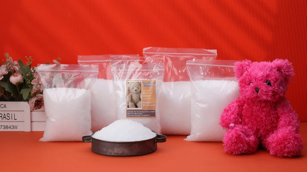 Multiple clear bags of white stuffing beads next to pink plush bear on orange surface.