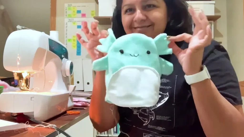 A woman proudly holds up a completed handmade plush toy, showing her sewing machine in the background.