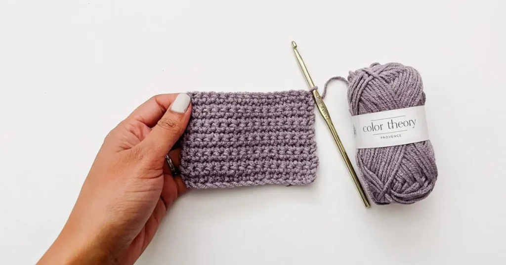 Hand holding a crochet swatch with purple yarn and hook.
