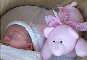 Sleeping baby next to soft pink teddy bear with checkered bow.