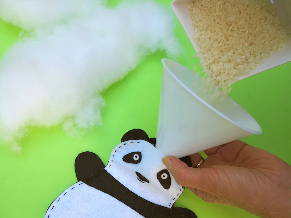 Hand pouring stuffing beads into panda plush using a funnel on green background.