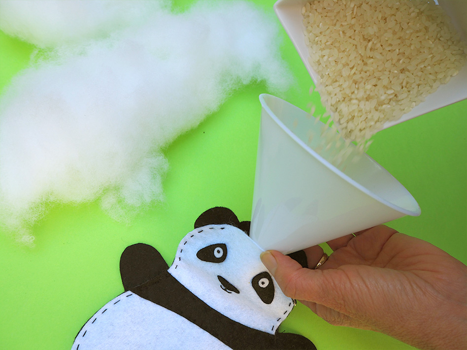 Hand using a funnel to fill a panda-shaped fabric toy with rice; fiberfill is visible nearby on a green surface